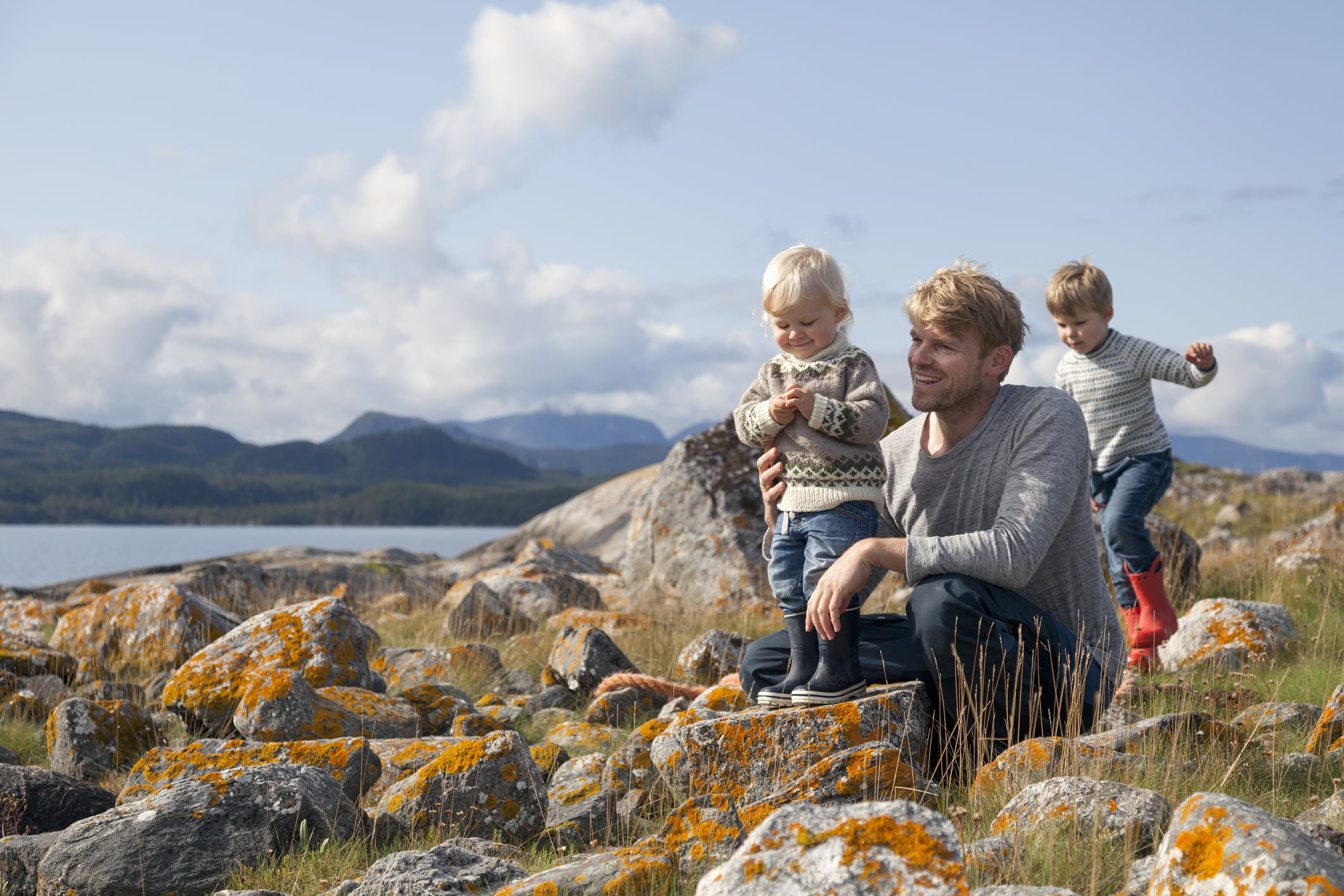 Far og to små barn nyter en dag ute i naturen, sitter på steiner dekket av lyng på en solfylt dag.