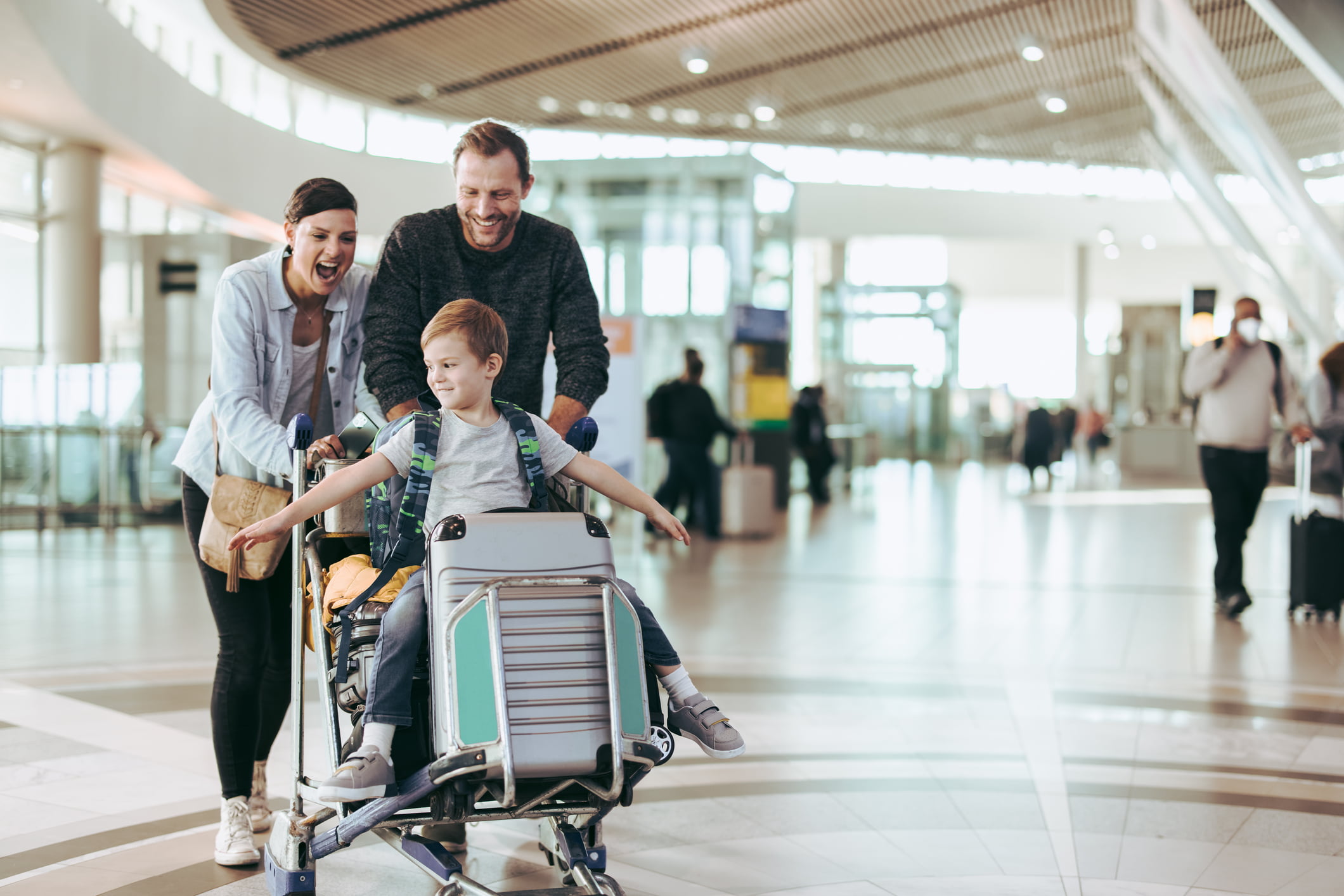 Familie på flyplassen ler og har det gøy, med et barn som sitter på en bagasjetralle.