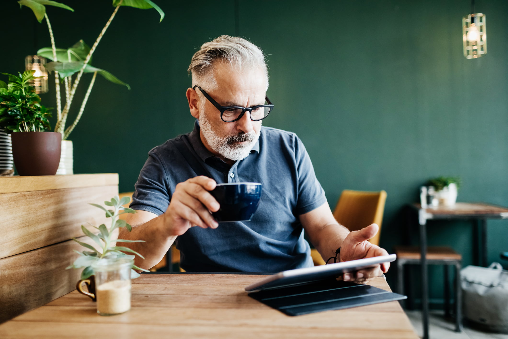 En eldre man drikker kaffe og leser noe på nettbrettet sitt med alvorlig ansiktsuttrykk.