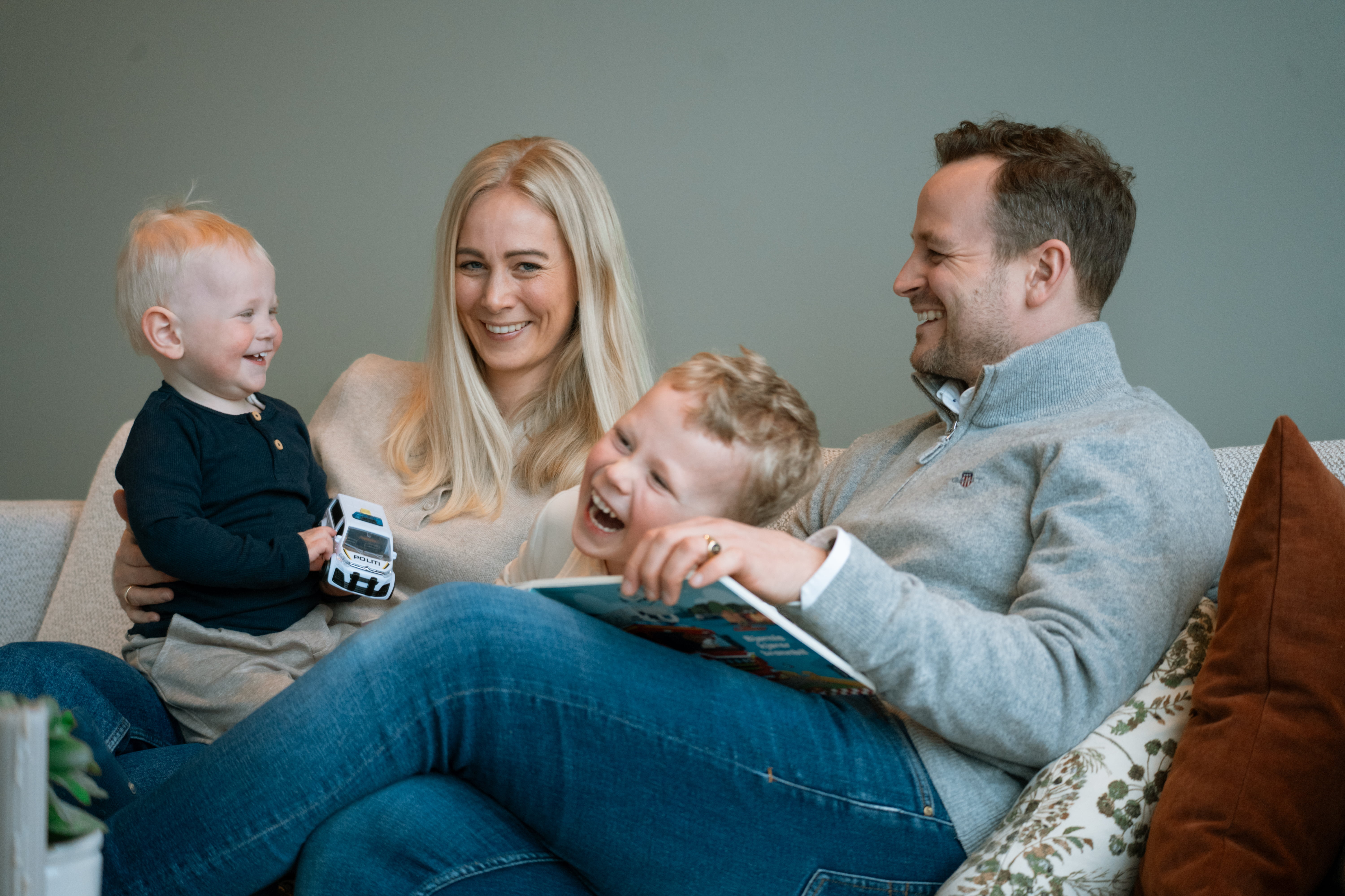 Familie sitter i sofaen og leser en bok sammen. Ett barn holder en lekebil, og det er puter og grønne planter i rommet.