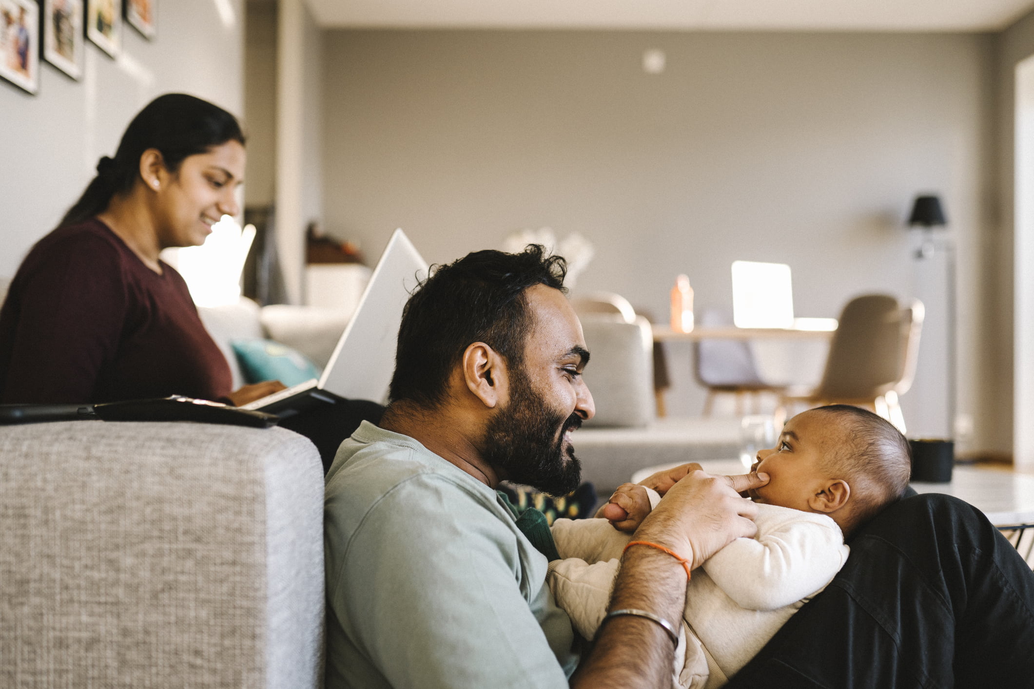 Familie på tre der mor sitter i sofa på pc, mens far sitter på gulvet og leker med baby