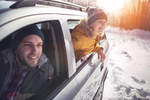 To smilende personer nyter vinterturen i en bil, en mann som kjører og et barn som lener seg ut av vinduet, begge kledd i varme luer, med en snødekt skog i bakgrunnen.